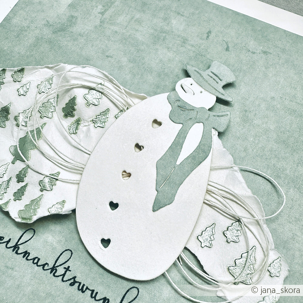 Mit Alexandra Renke Motivstanze Schneemann gestalteter Papierschneemann mit Schal und Mütze auf strukturiertem Designpapier mit Weihnachtsbaummustern und Schnurakzenten.