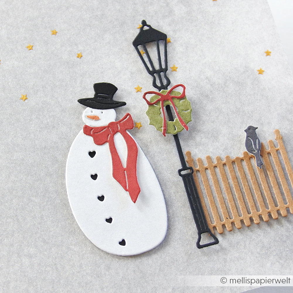 Papierschneemann mit rotem Schal an einem Laternenpfahl und Zaun, hergestellt mit Alexandra Renke Designpapiere und Motivstanze Schneemann, mit einem Vogel und goldenen Sternen auf hellem Hintergrund.