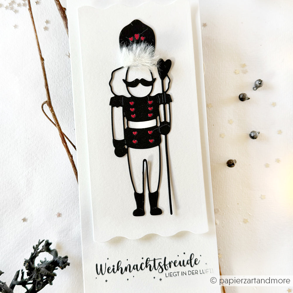 Handgefertigte Karte von Alexandra Renke mit einem schwarz-weißen Nussknacker-Motiv, das mit Designpapieren und den Lieblings-Nussknacker-Dies gestaltet wurde, akzentuiert mit roten Herzen und einem deutschen Weihnachtsgruß.