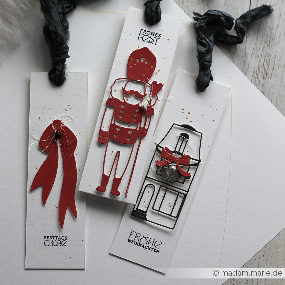 Drei handgefertigte weihnachtliche Lesezeichen von Alexandra Renke mit festlichen Motiven - eine rote Schleife, Motivstanze Lieblings-Nussknacker und ein geschmückter Kamin - hergestellt mit hochwertigen Designpapieren für einen charmanten Weihnachtslook.
