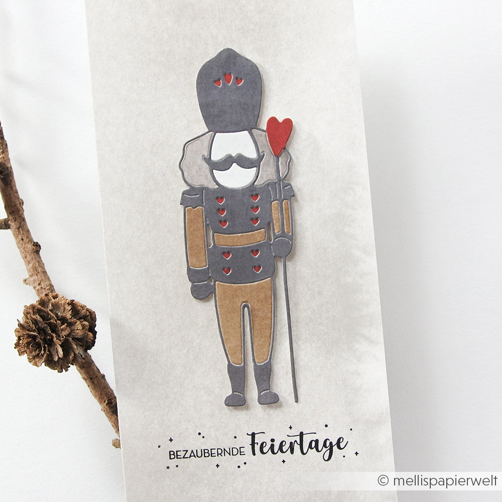 Ein Scherenschnitt von Alexandra Renke zeigt einen Lieblings-Nussknacker, der ein Herz in der Hand hält, darunter der Text "Bezaubernde Feiertage".