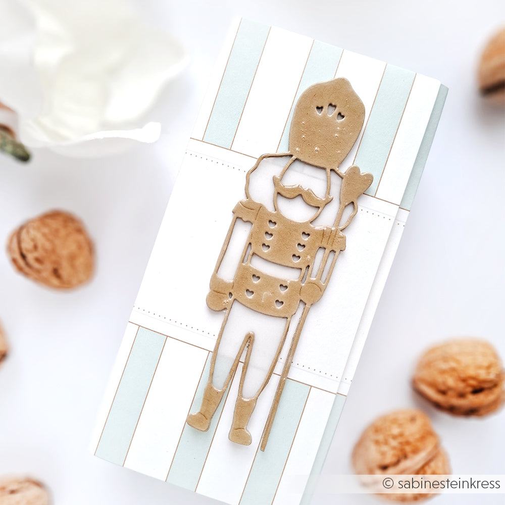 Eine Nussknacker-Dekoration aus Pappe, die mit Alexandra Renke Designpapieren und einer Motivstanze Nussknacker hergestellt wurde, sitzt auf einer gestreiften Geschenkbox, die von Walnüssen umgeben ist.