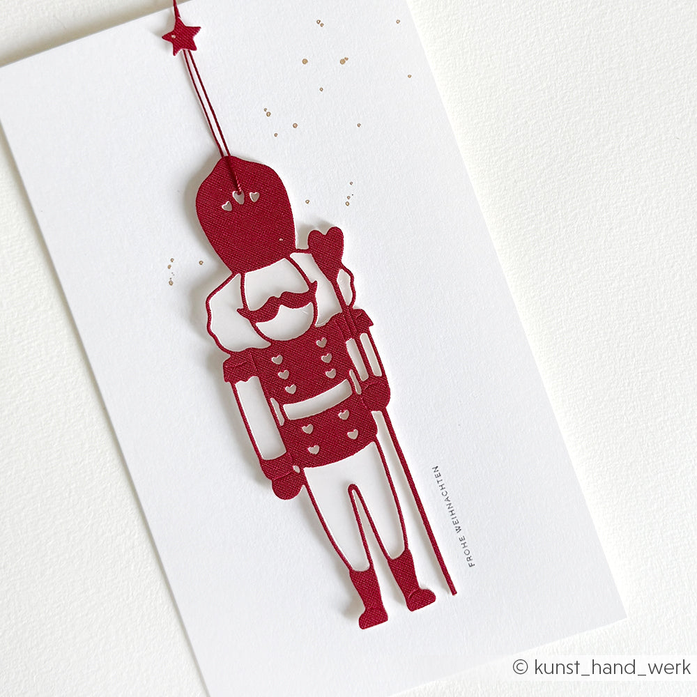 Festliche weiße Karte mit einem roten Nussknacker-Soldaten, der einen Luftballon mit goldenen Punkten hält. Gestaltet mit Alexandra Renke Motivstanze Lieblings-Nussknacker und Premium Designpapieren für ein einzigartiges DieCut Systeme Design.