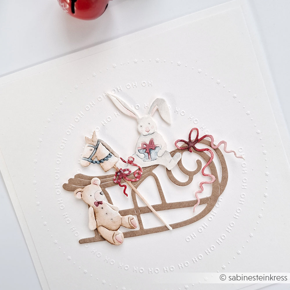 Eine Karte von Alexandra Renke zeigt einen Bären, einen Hasen und ein Schaukelpferd auf einem Schlitten mit geprägtem "Ho Ho Ho"-Text, hergestellt mit der Motivstanze Großer Schlitten für präzise Details.