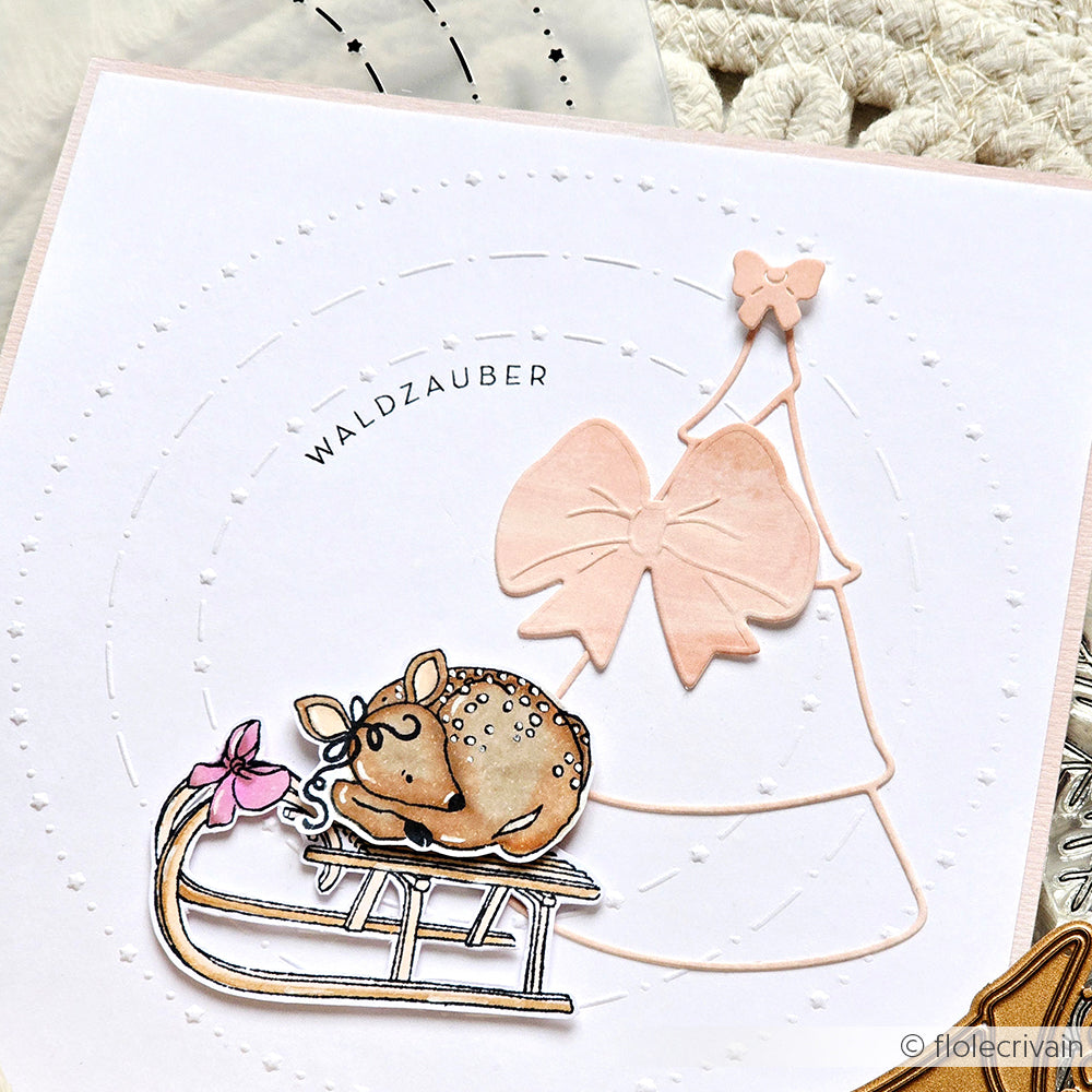 Handgefertigte Karte von Alexandra Renke mit einem Hirsch auf einem Schlitten, einer rosa Schleife und "WALDZAUBER" auf weißem Hintergrund mit gestanzten Stanze-Details für einen festlichen Touch.