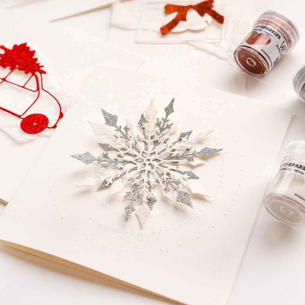 Eine handgefertigte Weihnachtskarte von Alexandra Renke zeigt eine silberne Glitzerschneeflocke, die mit DieCut Systemen und der Motivstanze Große Schneeflocke hergestellt wurde, umgeben von festlichem Bastelmaterial.