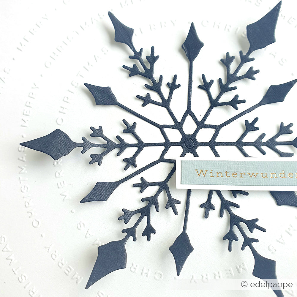 Eine dunkelblaue Papierschneeflocke auf weißem Grund mit der Aufschrift "Winterwunder", hergestellt mit der Motivstanze Große Schneeflocke von Alexandra Renke - ein schwacher, kreisförmiger Text, ideal für kreative Stanzsysteme oder Designpapiere.