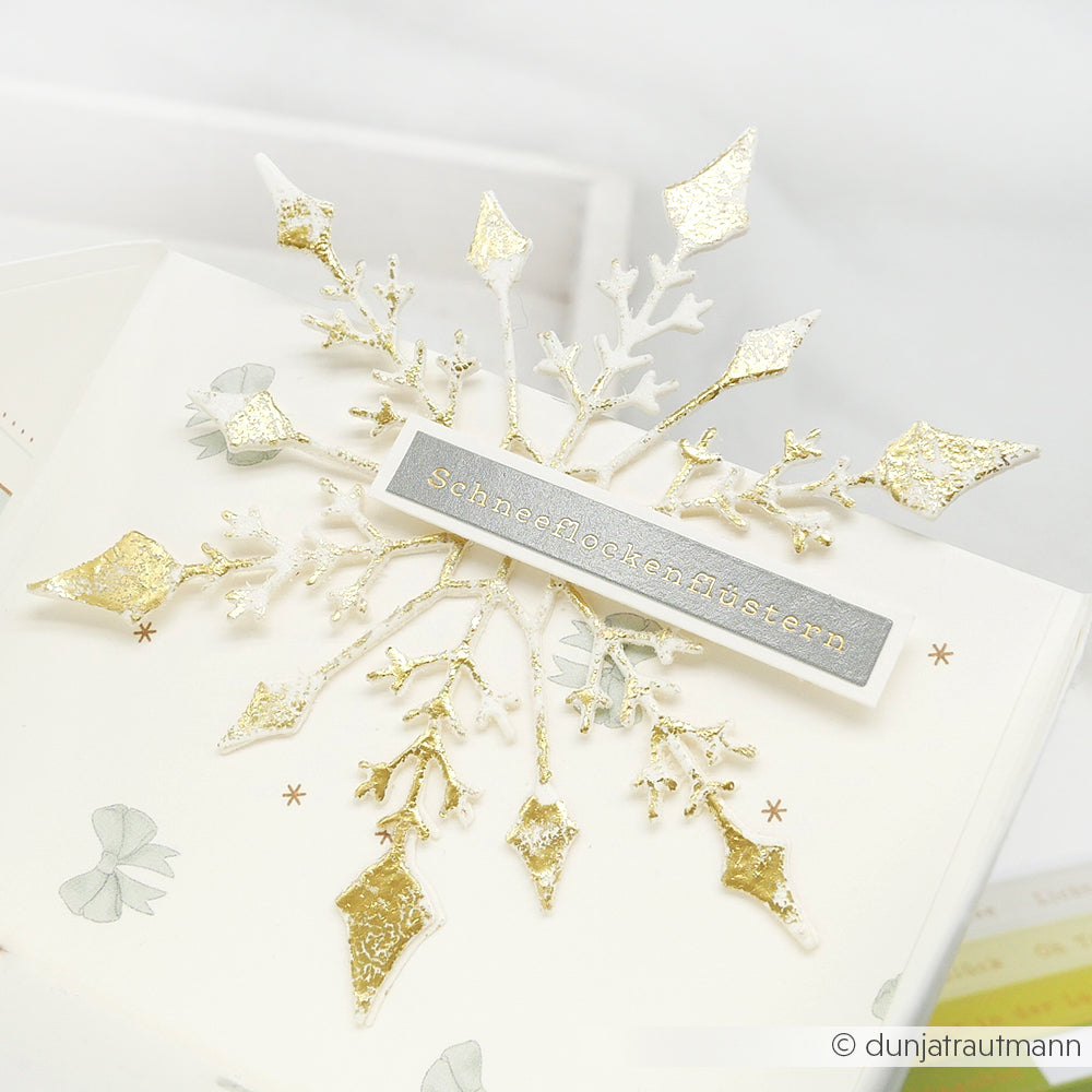 Gold-weiß glitzernde Schneeflockenkarte mit "Schneeflockenflüstern" in der Mitte, gestaltet mit der Motivstanze Große Schneeflocke von Alexandra Renke - ideal für alle Liebhaber von Stanzen für Papier.