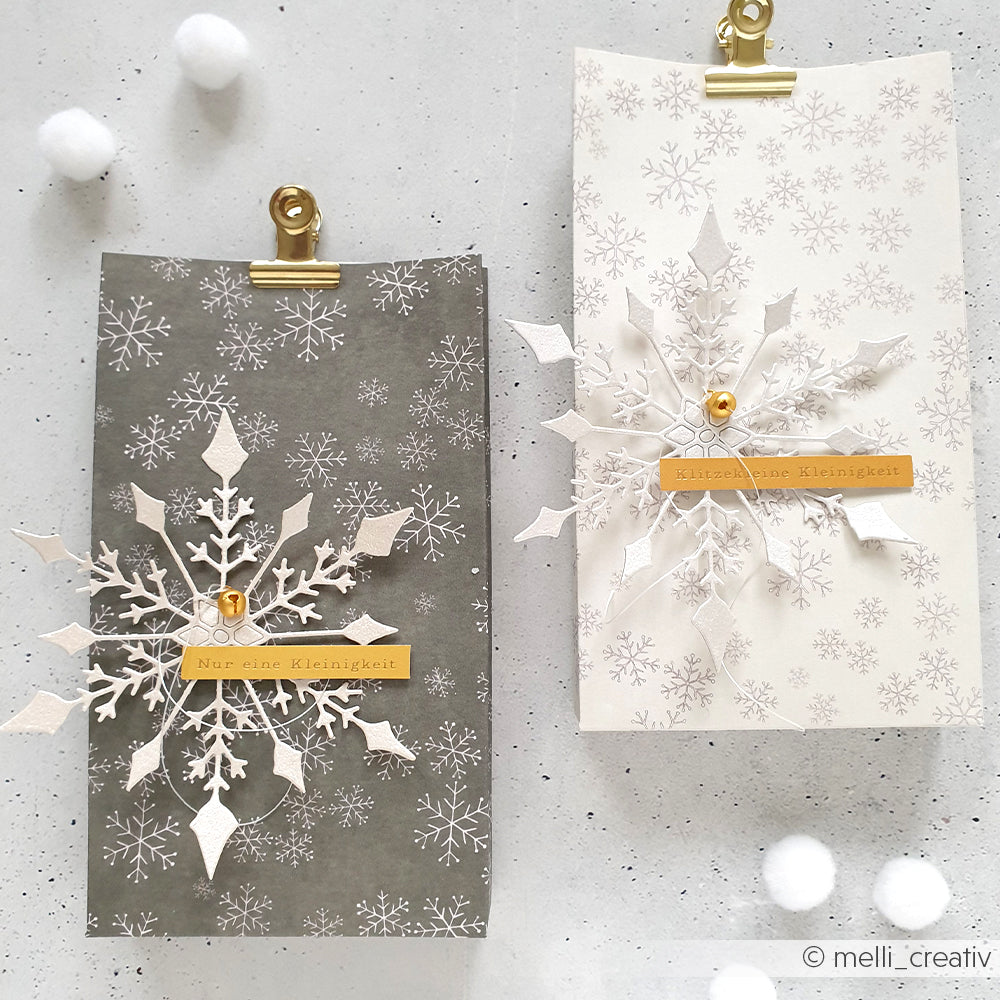 Zwei Geschenktüten mit Schneeflockenmotiven, hergestellt mit Alexandra Renke Motivstanze Große Schneeflocke Dies - eine grau, eine weiß - verziert mit goldenen Clips und Etiketten. Ideal zum Verschenken oder für kreative Projekte mit Alexandra Renke Stanzsystemen.