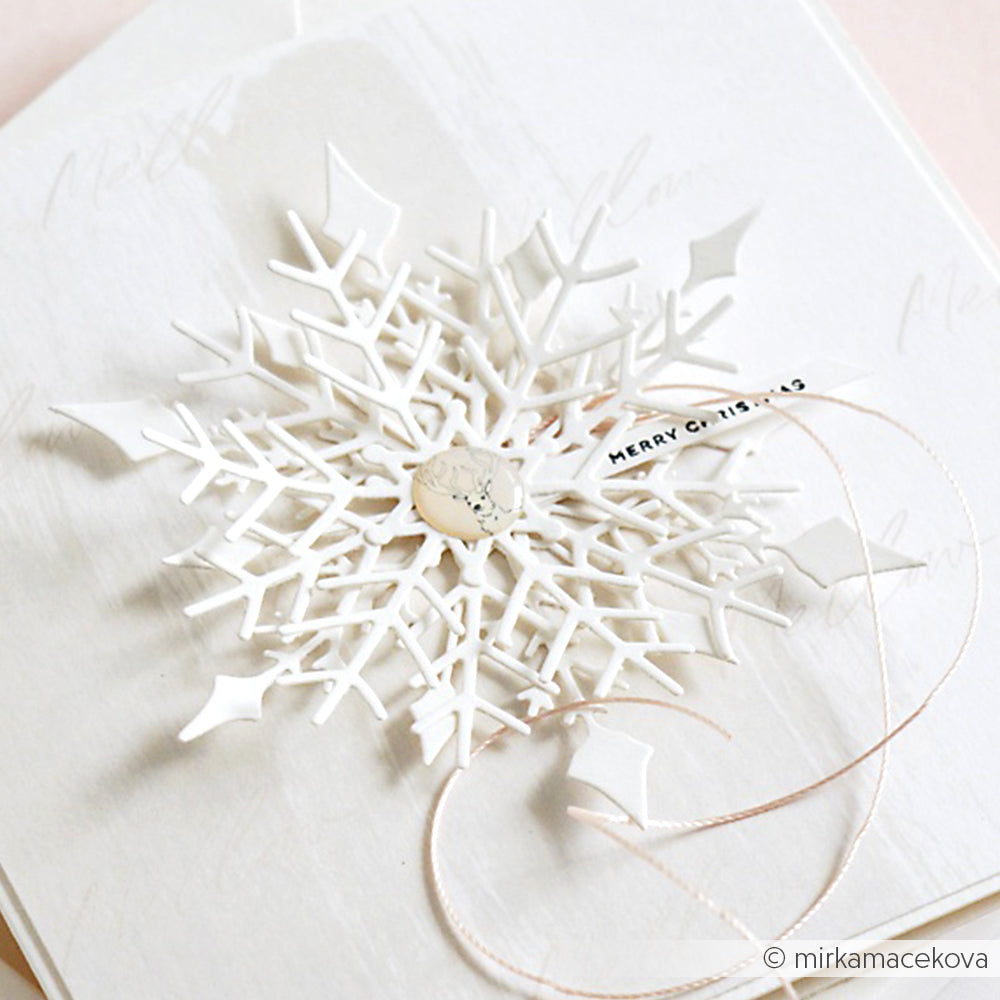 Die Schneeflocke aus weißem Papier, die mit der Motivstanze Große Schneeflocke von Alexandra Renke und hochwertigen Designpapieren hergestellt wurde, ist mit einem Knopf in der Mitte auf weißem Strukturkarton versehen und mit einer dünnen Schnur zum Aufhängen versehen.