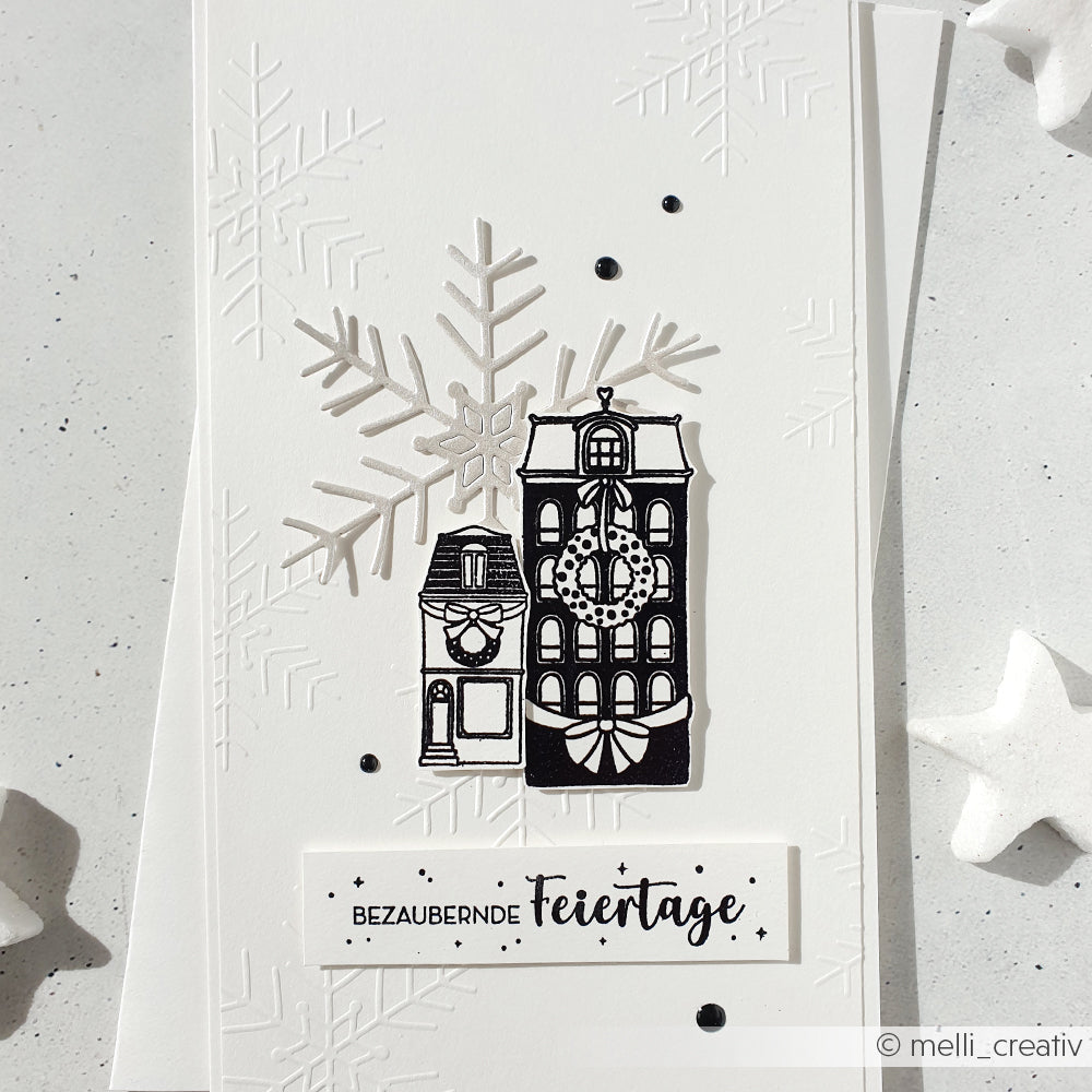 Eine weiße Weihnachtskarte von Alexandra Renke auf 200g Designpapieren zeigt schwarze Häuser, eine mit der Motivstanze Große Schneeflocke gestanzte Schneeflocke und den Gruß BEZAUBERNDE Feiertage.