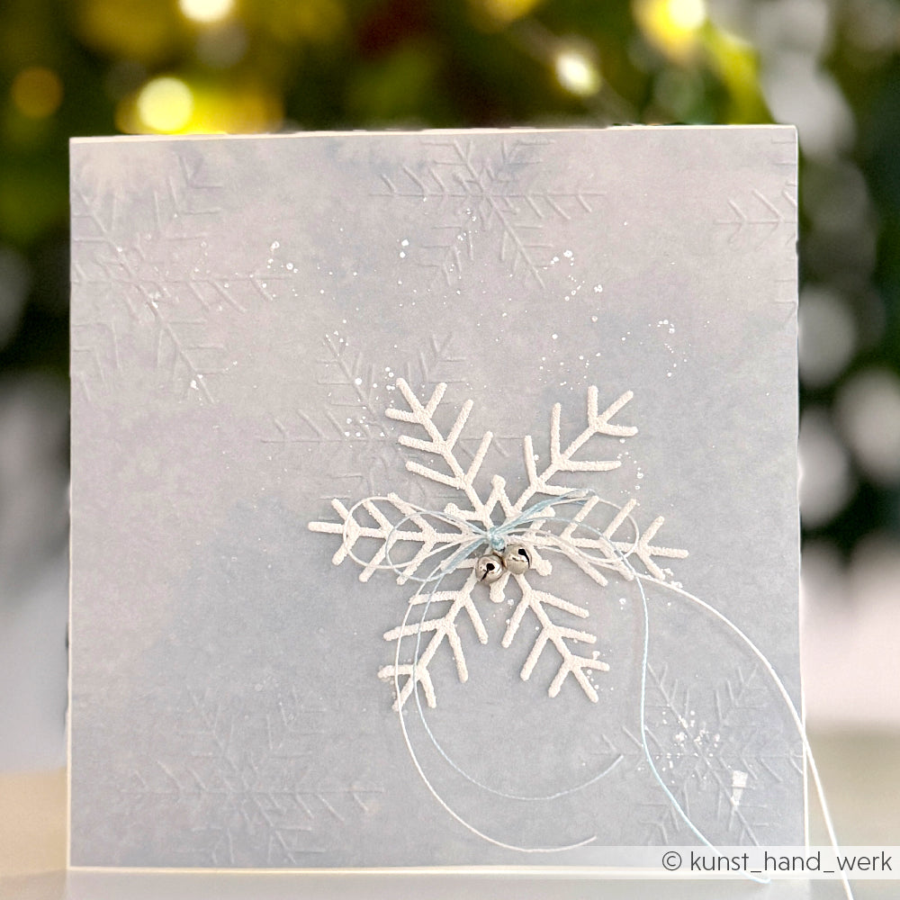 Quadratische Karte von Alexandra Renke mit geprägten Schneeflocken und einer weißen, glitzernden Schneeflocke, die mit der Motivstanze Große Schneeflocke gestanzt wurde und in der Mitte mit einer Schnur abgeschlossen ist.
