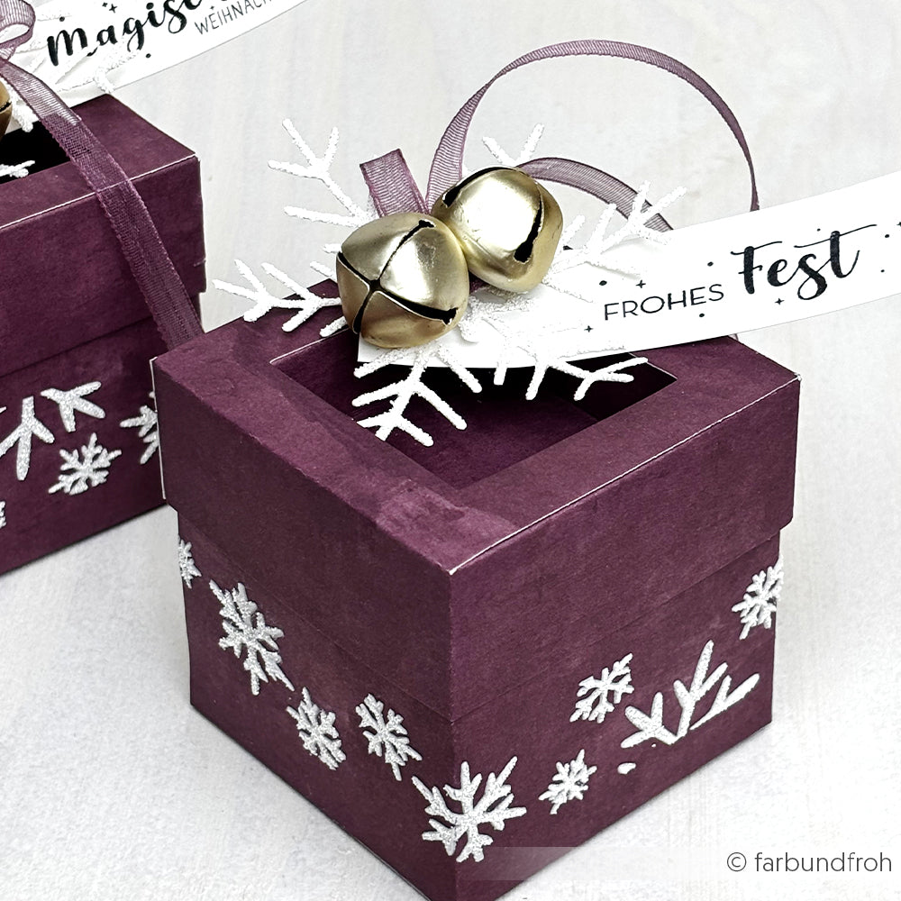 Lila Geschenkbox aus Alexandra Renke Designpapier, verziert mit weißen Schneeflocken, die mit der Großen Schneeflocke gestanzt wurden, silbernen Jingle Bells und einem "Frohes Fest"-Anhänger.