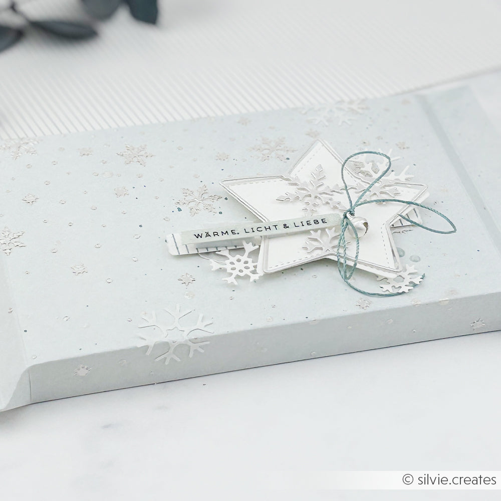 Geschenkbox mit einer Motivstanze in Form einer Schneeflocke und einem Stern-Anhänger mit der Aufschrift WÄRME, LICHT & LIEBE.
