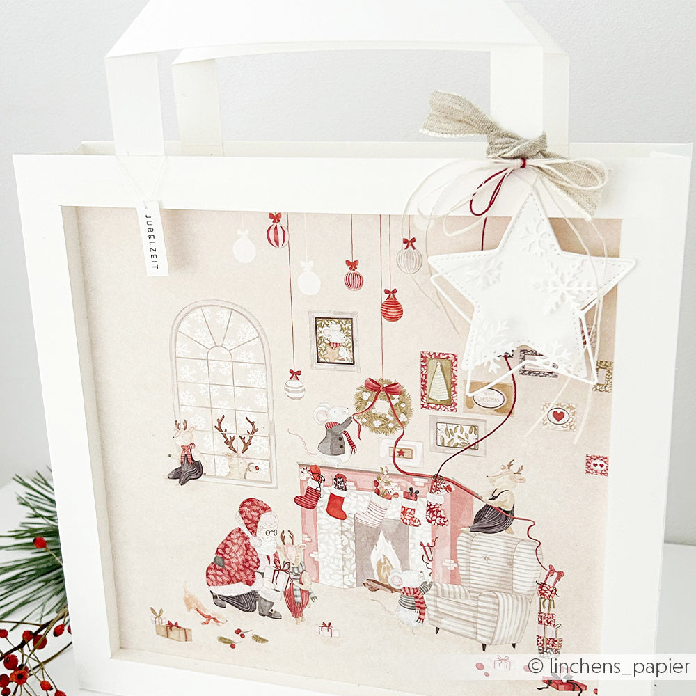 Eine weiße Geschenktüte mit einem Weihnachtsmotiv, auf dem der Weihnachtsmann und Mäuse abgebildet sind, verziert mit Designpapieren.