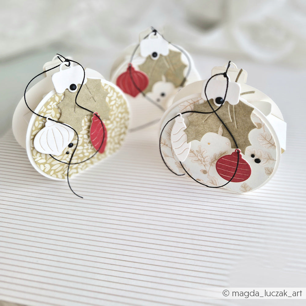 Drei runde Papierornamente mit winterlichem Blumenkonfetti, Stechpalmenzweigen und hängenden Kugeln.