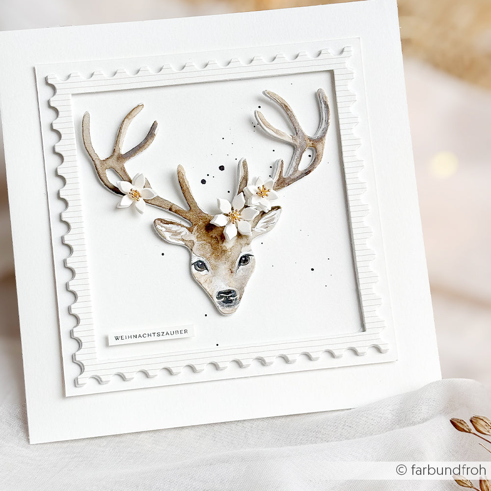 Handgefertigte Karte mit einem Hirschkopf, verziert mit weißen Blumen und winterlichem Blumenkonfetti, mit der deutschen Aufschrift „WEIHNACHTSZAUBER“.