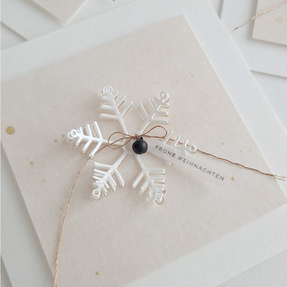 Ein weißes Schneeflocken-Ornament, das mit einer Motivstanze „Schneeflocke“ gestaltet wurde, auf einer Karte mit einem Anhänger, auf dem „FROHE WEIHNACHTEN“ steht.
