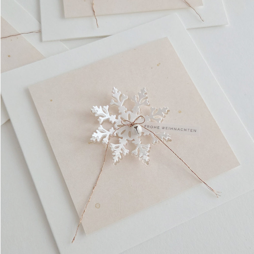 Ein weißes Schneeflocken-Ornament, das mit einer „Schneeflocke“-Motivstanze gestaltet und auf einer quadratischen Karte mit der Aufschrift „FROHE WEIHNACHTEN“ befestigt wurde.