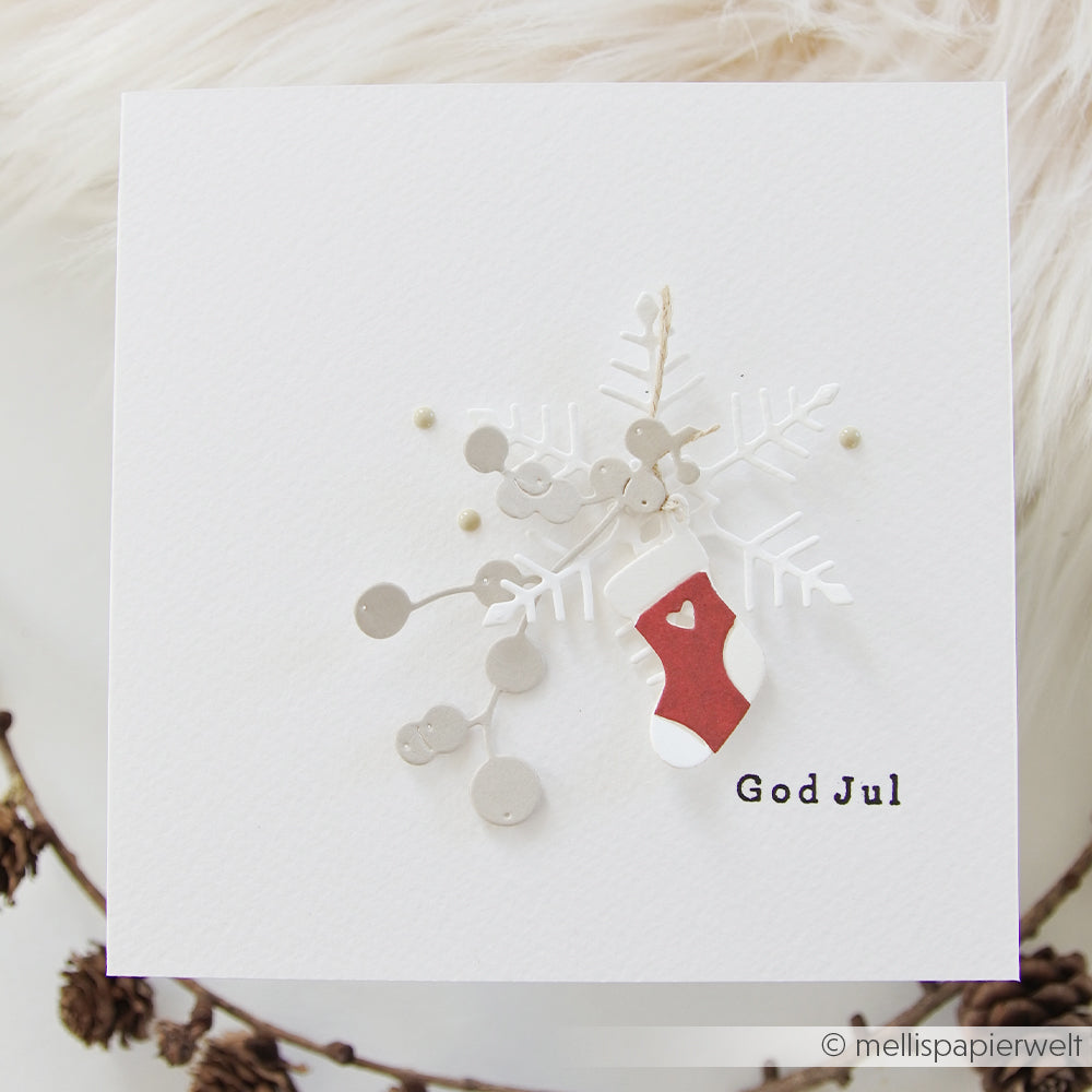 Eine quadratische Weihnachtskarte mit einer Papierschneeflocke, die mit der Motivstanze „Schneeflocke“ gestaltet wurde, Beeren und einem kleinen roten Strumpf mit der Aufschrift „God Jul“.