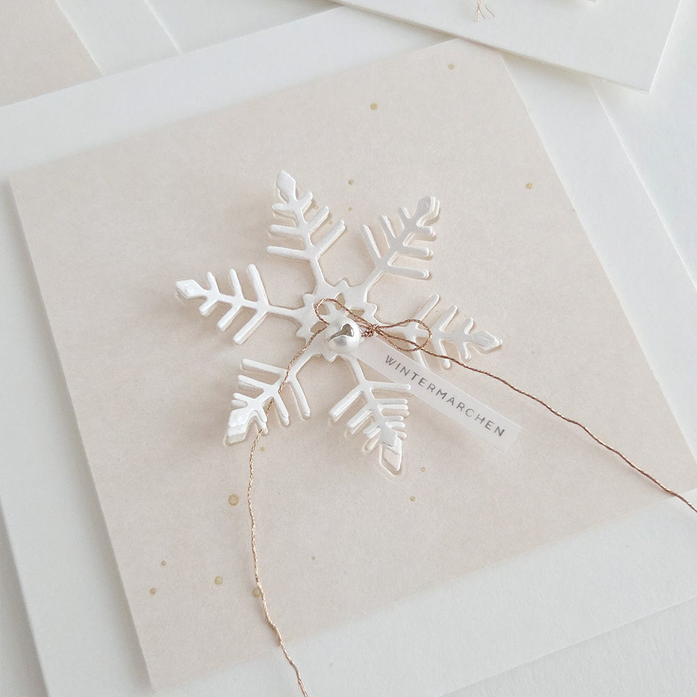 Ein weißes Keramik-Schneeflocken-Ornament, das mit einer Motivstanze „Schneeflocke“ hergestellt und mit einer Schnur an einer „Wintermärchen“-Grußkarte befestigt wurde.