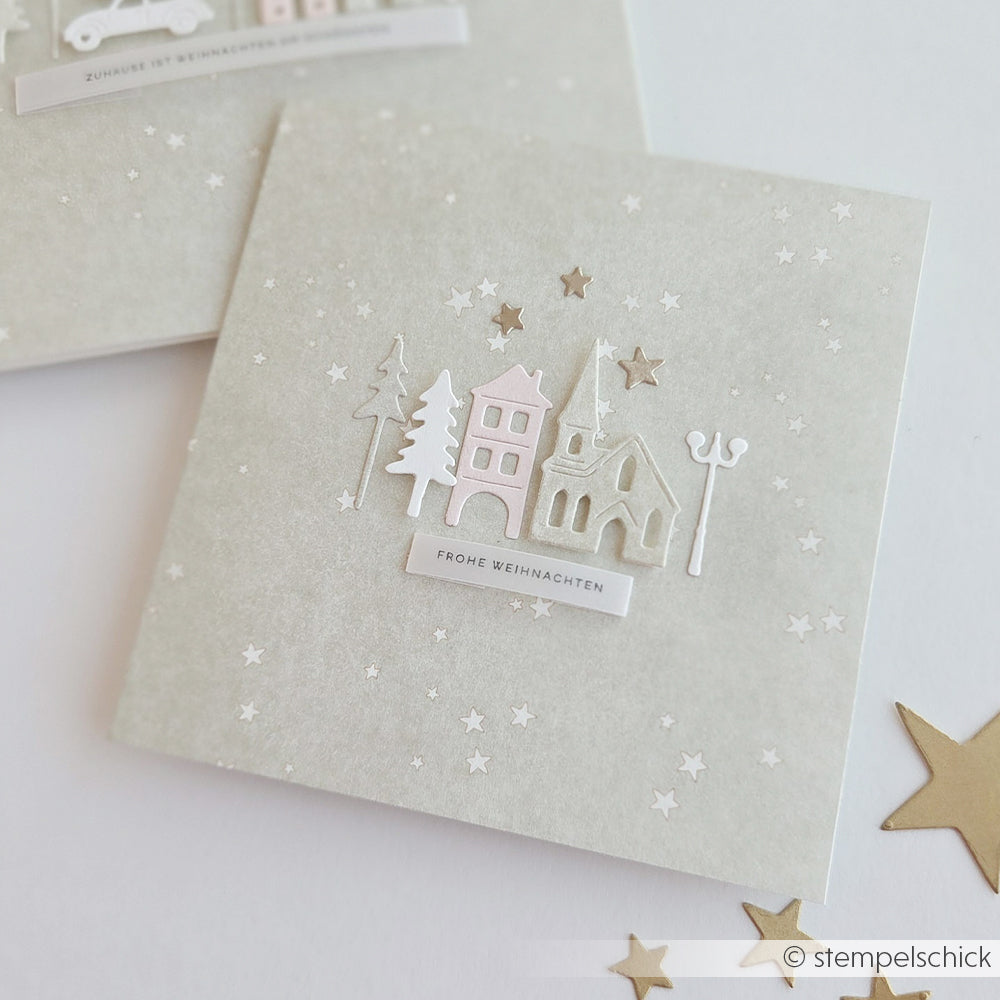 Eine quadratische, handgefertigte Weihnachtskarte mit einer Papierdorfszene, die aus Motiven eines Stanzsystems gestaltet wurde, sowie dem Schriftzug „Frohe Weihnachten“.