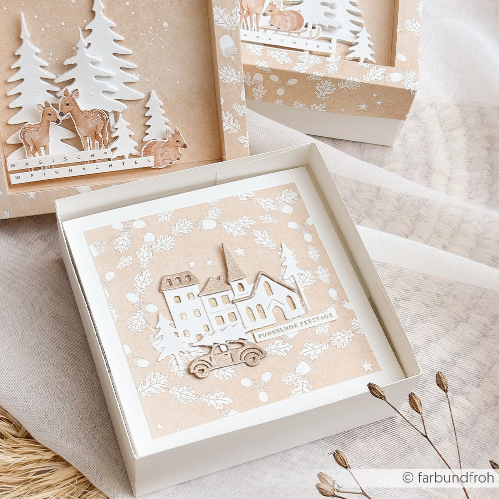 Weiße Geschenkbox mit einer Weihnachtskarte, die mit einer Motivstanze mit Weihnachtsdorf-Motiv gestaltet wurde.