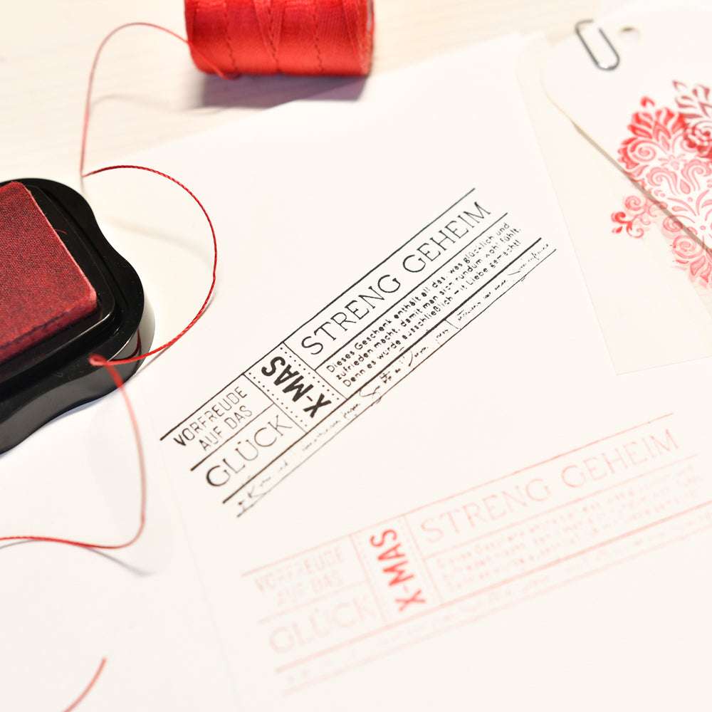 Ein gestempeltes Streng Geheim XMAS-Etikett von Alexandra Renke erscheint auf weißem Papier, hergestellt mit einem Holzstempel. Roter Faden und ein Stempelkissen liegen daneben - ideal für Papierprojekte.