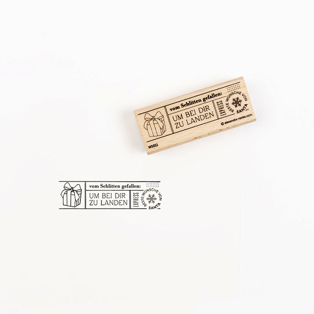 Ein Holzstempel, der eine Geschenkbox und einen deutschen Text neben dem Stempelabdruck zeigt.
