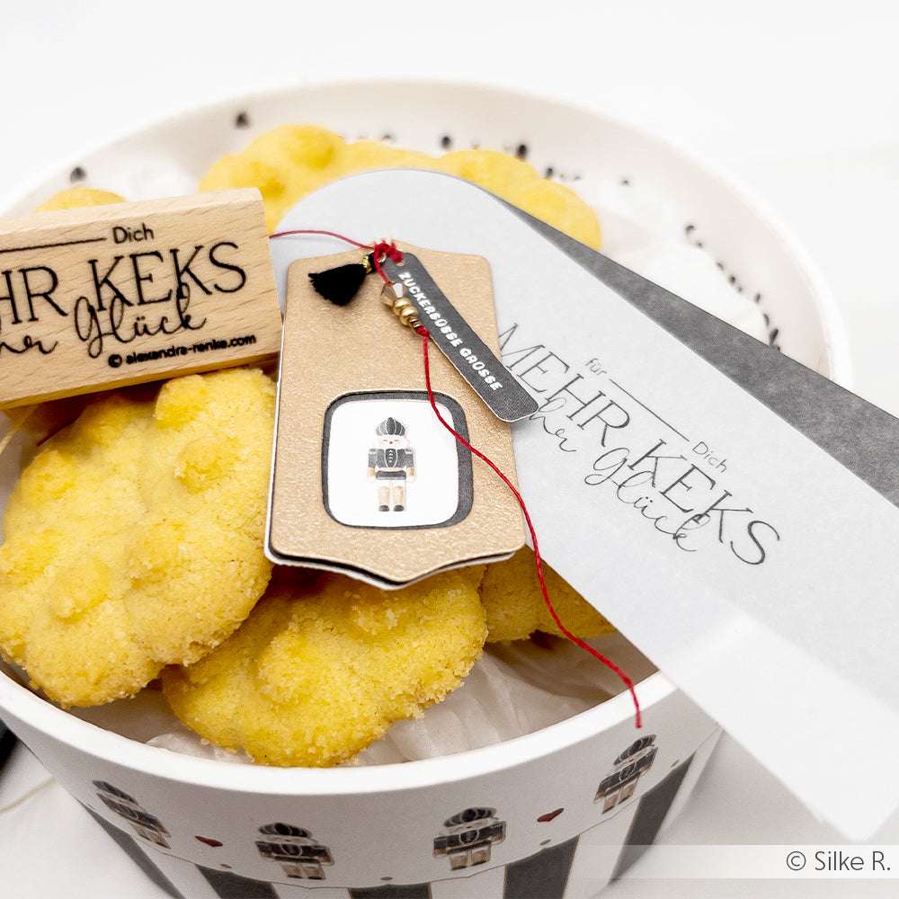 Alexandra Renke runde Geschenkbox mit gelben Keksen, einem Holzstempel und einem MEHR KEKS Glück Anhänger auf weißem Hintergrund.
