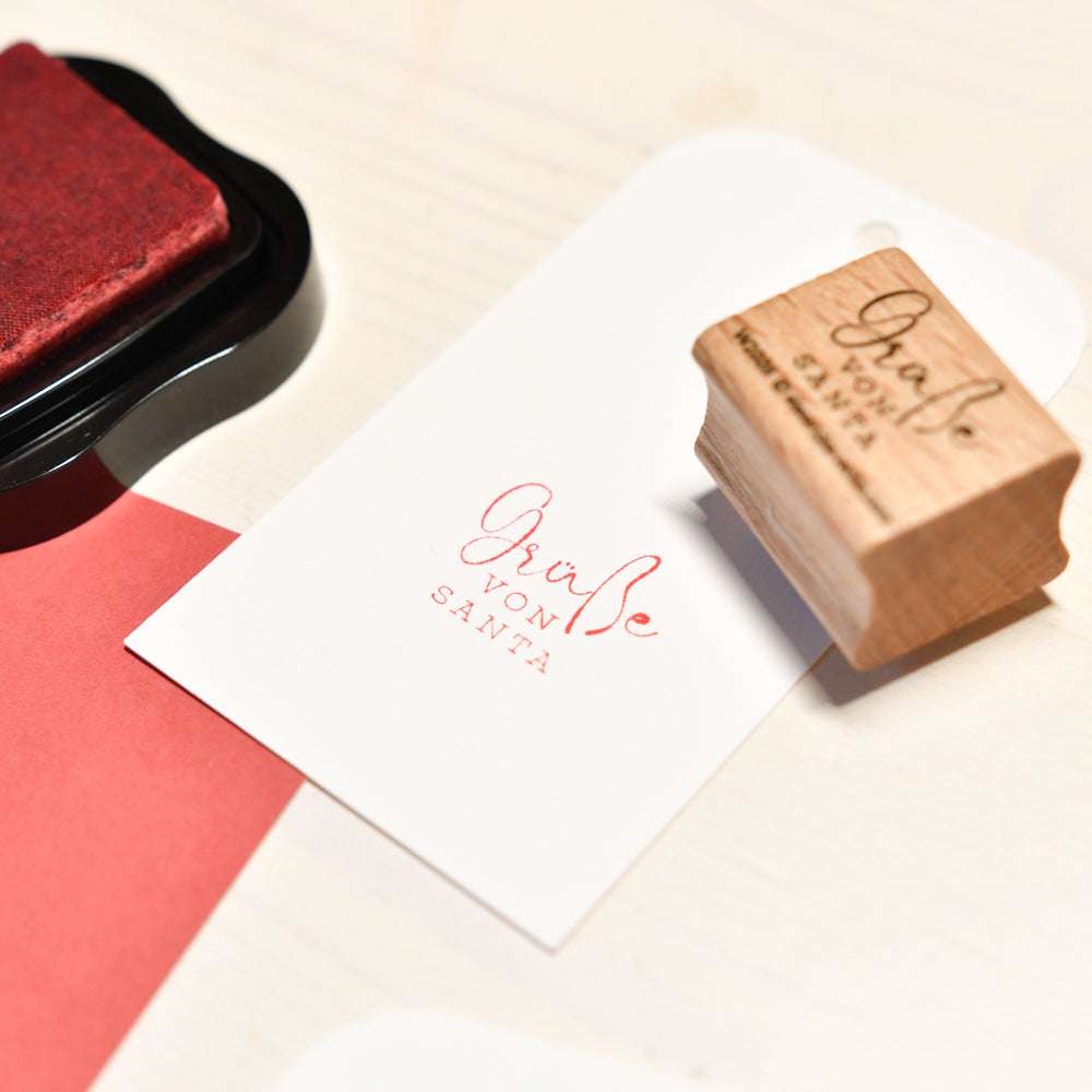 Ein Holzstempel und ein Stempelkissen von Alexandra Renke liegen neben einem weißen Tag, der mit "Grüße von Santa" in roter Tinte bestempelt wurde - ideal für festliche Papierprojekte.