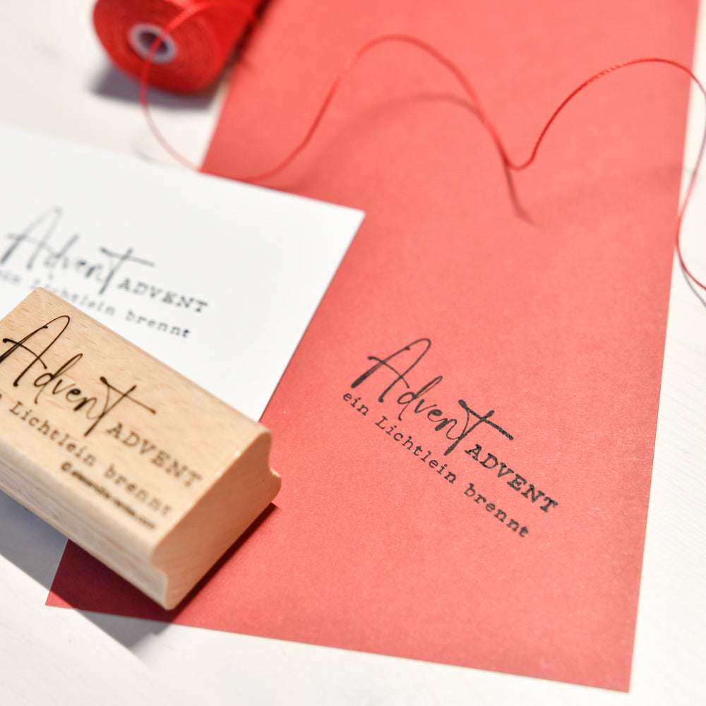 Ein Holzstempel von Alexandra Renke mit "Advent Advent ein Lichtlein brennt", sowie rotes Papier und Garn für Ihre Papierprojekte.