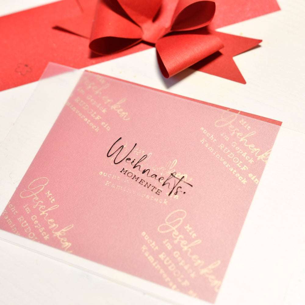 Rosa Karte mit "Weihnachtsmomente"-Text, gestempelt mit Stempelgummi von Alexandra Renke, neben einer roten Schleife und einem Umschlag auf weißem Untergrund.