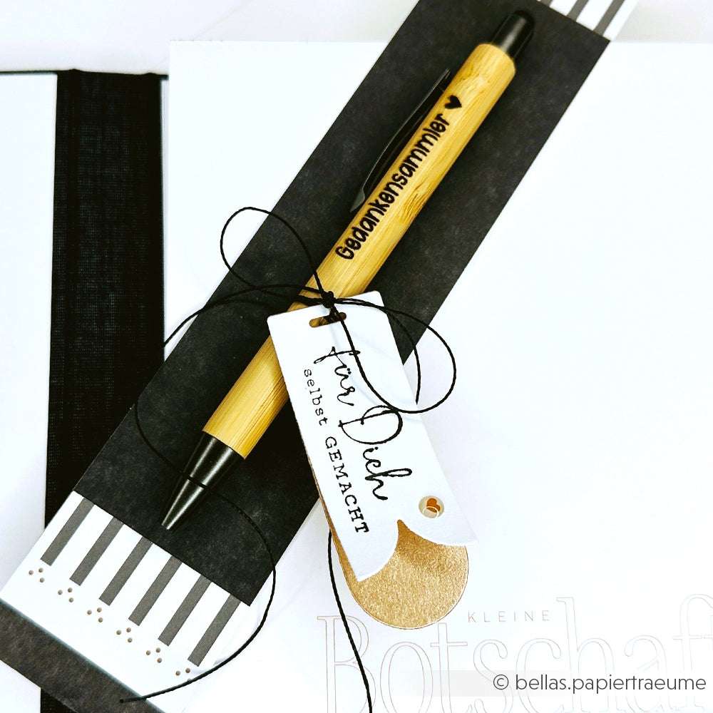Ein Alexandra Renke Bambus Kugelschreiber mit Gedankenschmiedler, einem Holzstempel und einem "Für Dich selbst gemacht" Geschenkanhänger auf einem gestreiften Notizbuch.