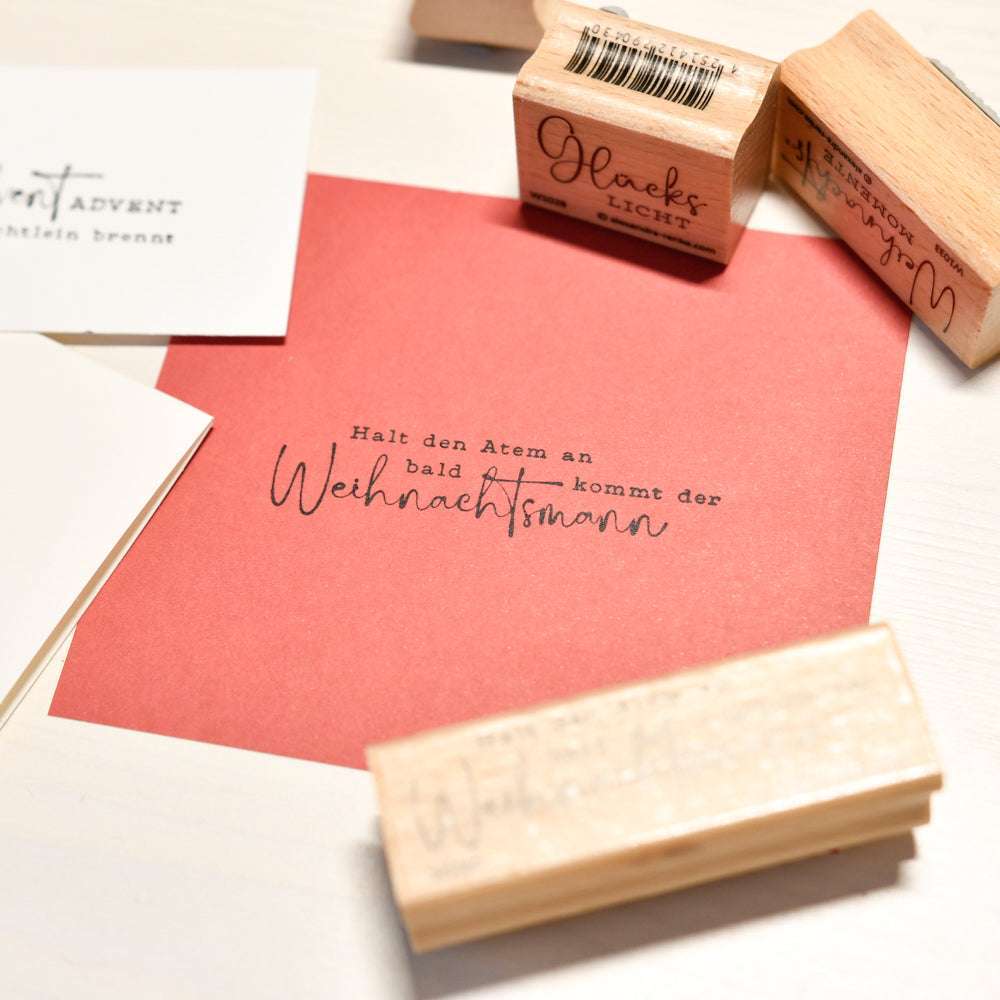 Rote Karte mit deutschem Text von Alexandra Renke, umgeben von Weihnachtsmann Stempel, Stempelkissen und weißen Karten auf einer hellen Oberfläche.