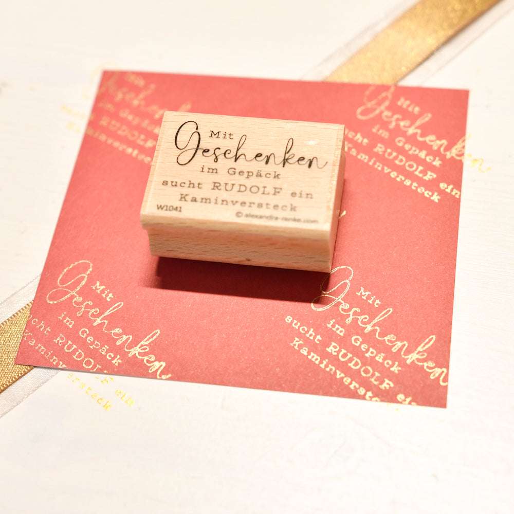 Ein Holzstempel von Alexandra Renke auf rotem Papier zeigt deutschen Text zu Geschenken und Rudolph dem Rentier - ideal für kreative Papierprojekte mit passendem Stempelkissen.