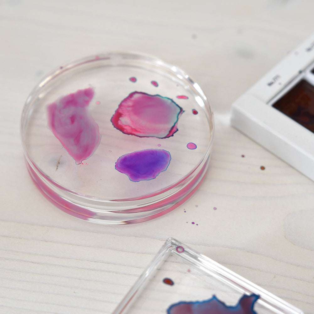 Eine Petrischale mit rosafarbenen und violetten Aquarellflecken steht auf einer hellen Holzfläche neben einem Alexandra Renke Acryl Stempelblock (0 cm x 8 cm).
