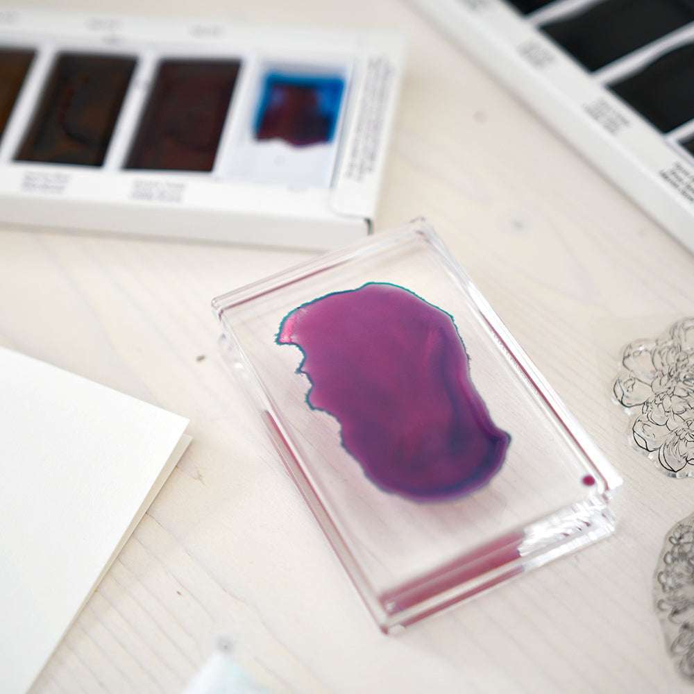 Ein lilafarbener Tintenklecks auf einem Alexandra Renke Stempelblock 5, einem durchsichtigen, rechteckigen Acrylblock für die präzise Positionierung von Stempeln, liegt auf einem Schreibtisch mit Kunstzubehör in der Nähe.