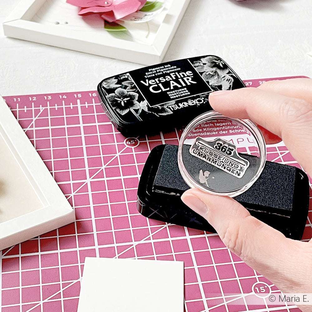 Eine Hand drückt einen Alexandra Renke Acryl Stempelblock auf ein schwarzes Stempelkissen und positioniert den Stempel vorsichtig auf einer rosafarbenen Bastelmatte, umgeben von anderen Bastelmaterialien.