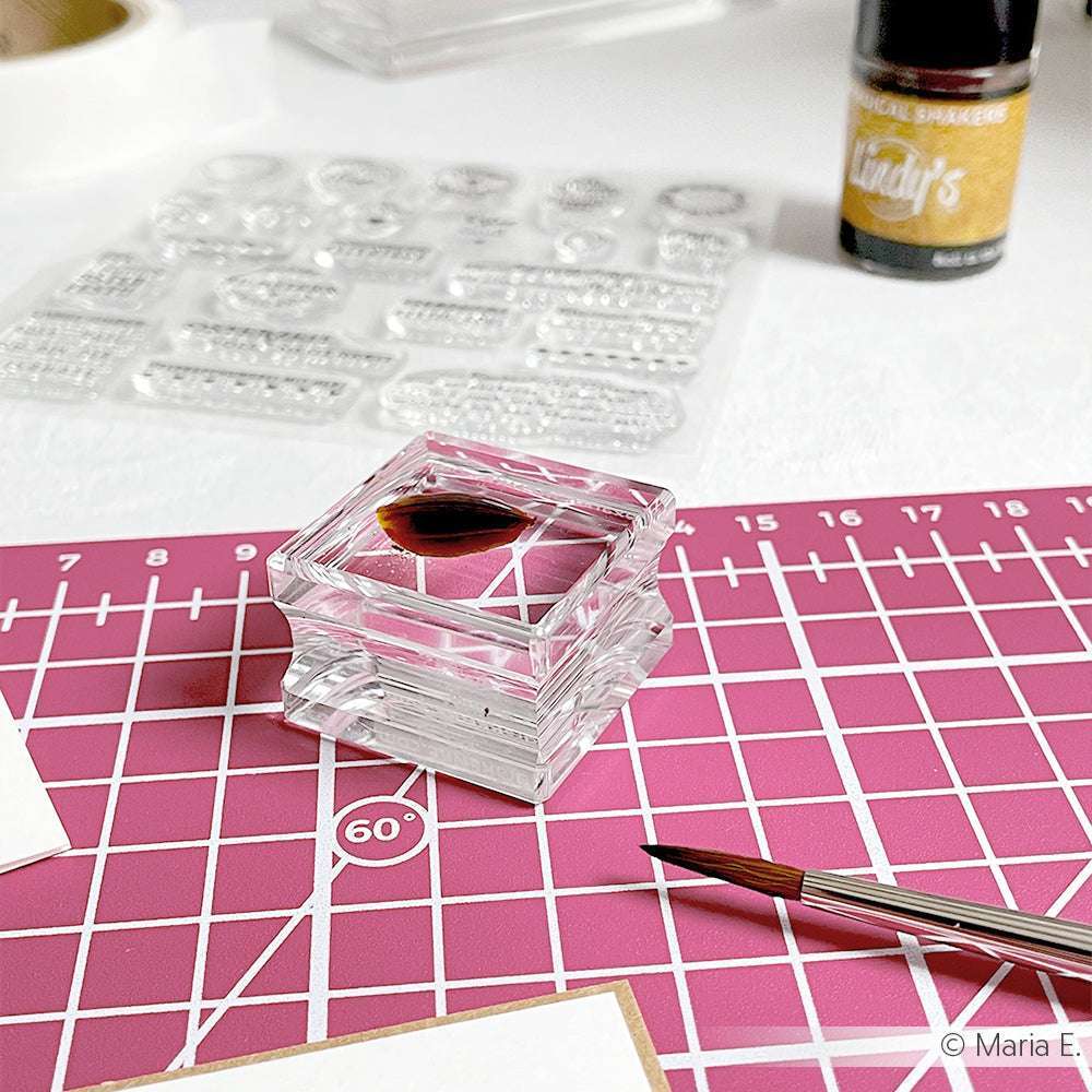 Der Alexandra Renke Clear Acryl Stempelblock (3,0 cm x 3 cm) zeigt rote flüssige Akzente auf einer rosa Schneidematte, mit einem Pinsel und verschiedenen Bastelmaterialien im Hintergrund.