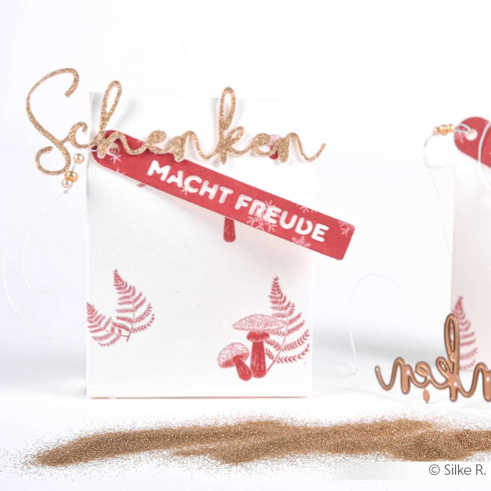 Weiße Geschenktüte mit goldenem "Schenken"-Schriftzug, rotem "Macht Freude"-Etikett, Farn- und Pilzmotiven und goldenem Glitzerschimmer, hergestellt mit WOW Embossing Powder.