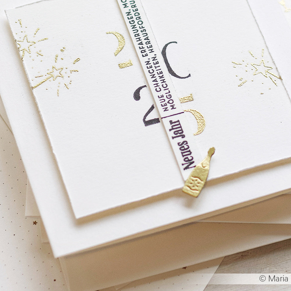 Ein Stapel weißer Karten, verziert mit goldenen Sternen, die mit WOW Embossing Powder und einem Heißprägewerkzeug hergestellt wurden, mit einer teilweise enthüllten deutschen Neujahrsbotschaft und goldenem Konfetti.