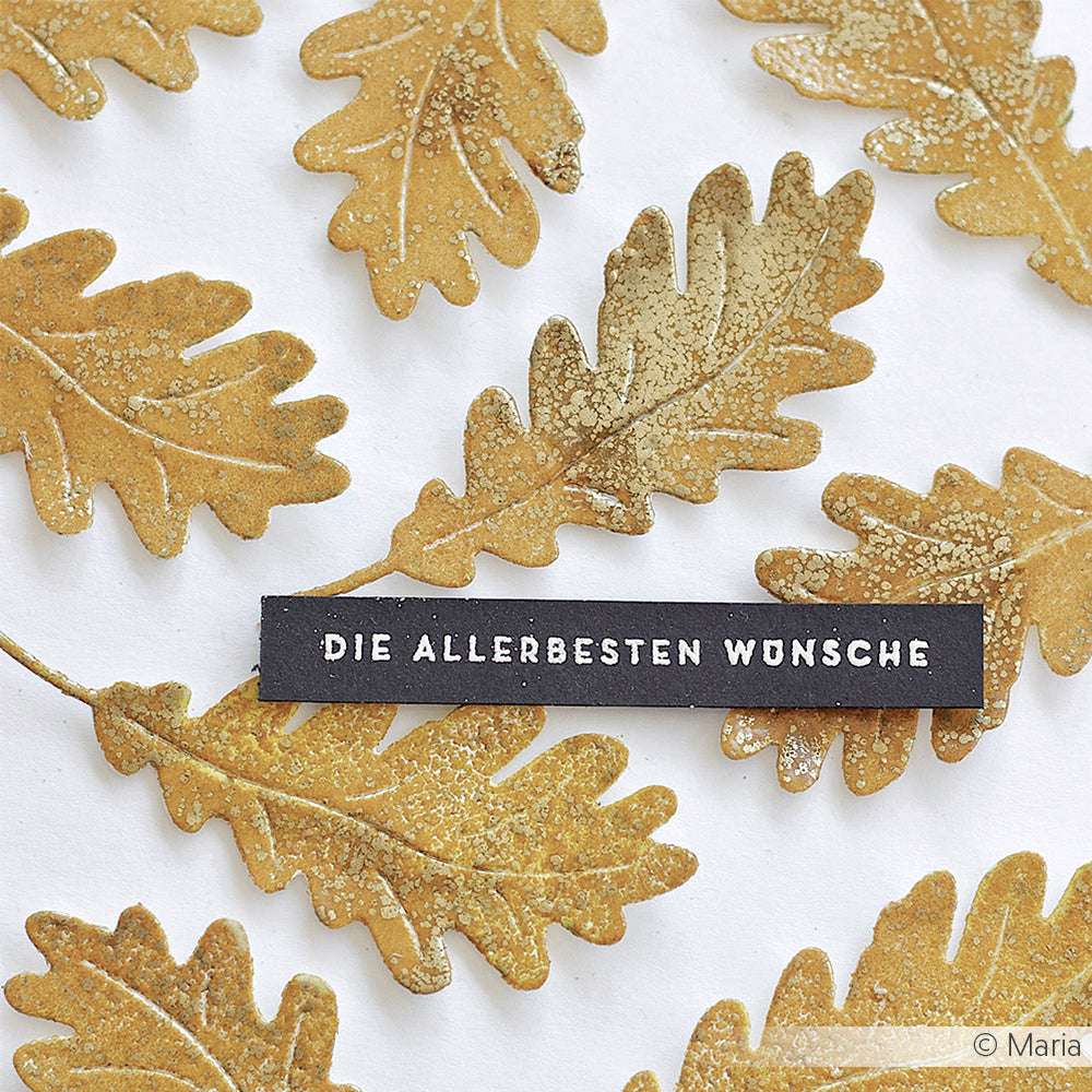 Goldenes Eichenlaub umrahmt ein schwarzes Etikett mit weißem Text - DIE ALLERBESTEN WÜNSCHE -, der durch den Schimmer des WOW Embossing Powders und die feinen Details eines Embossing-Farbkissens hervorgehoben wird.