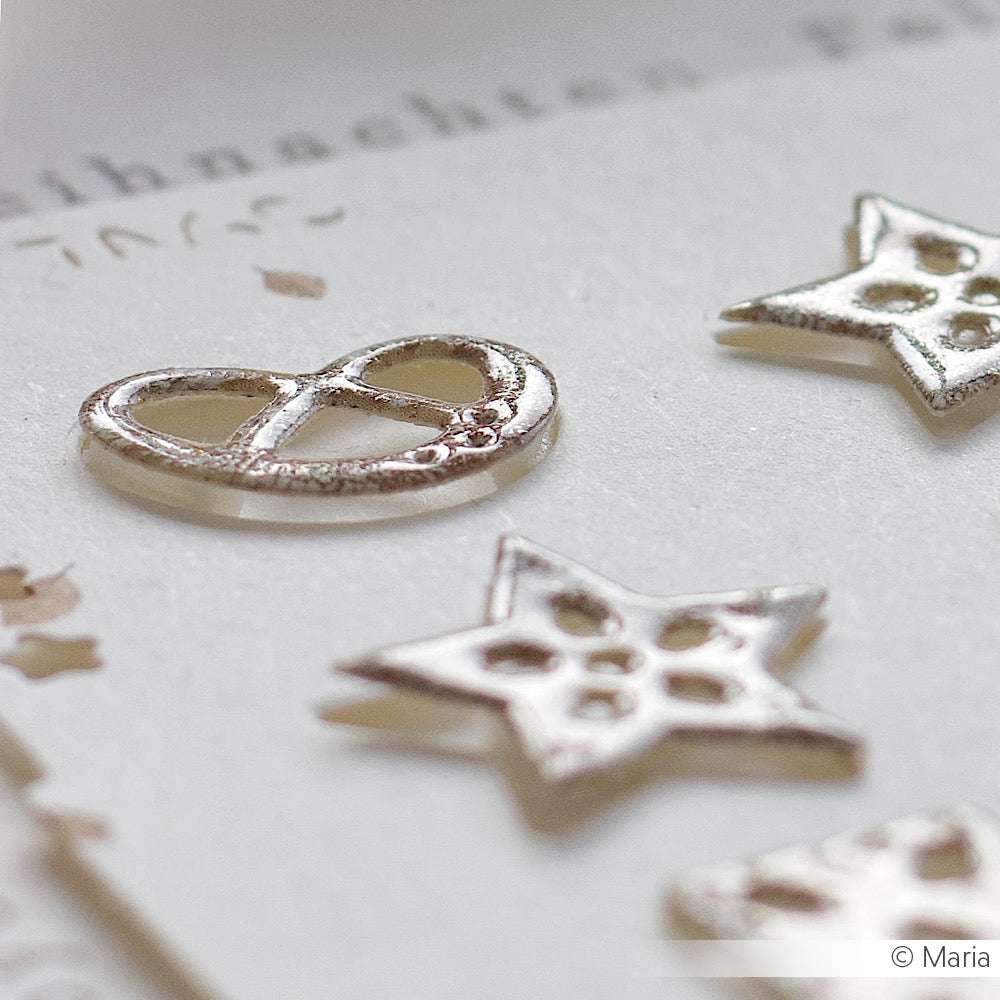 Silberne metallische Brezel- und Sternformen ruhen auf einem weißen, strukturierten Hintergrund. Ihr Glanz wird durch WOW Embossing Powder für einen atemberaubenden Prägeeffekt verstärkt.