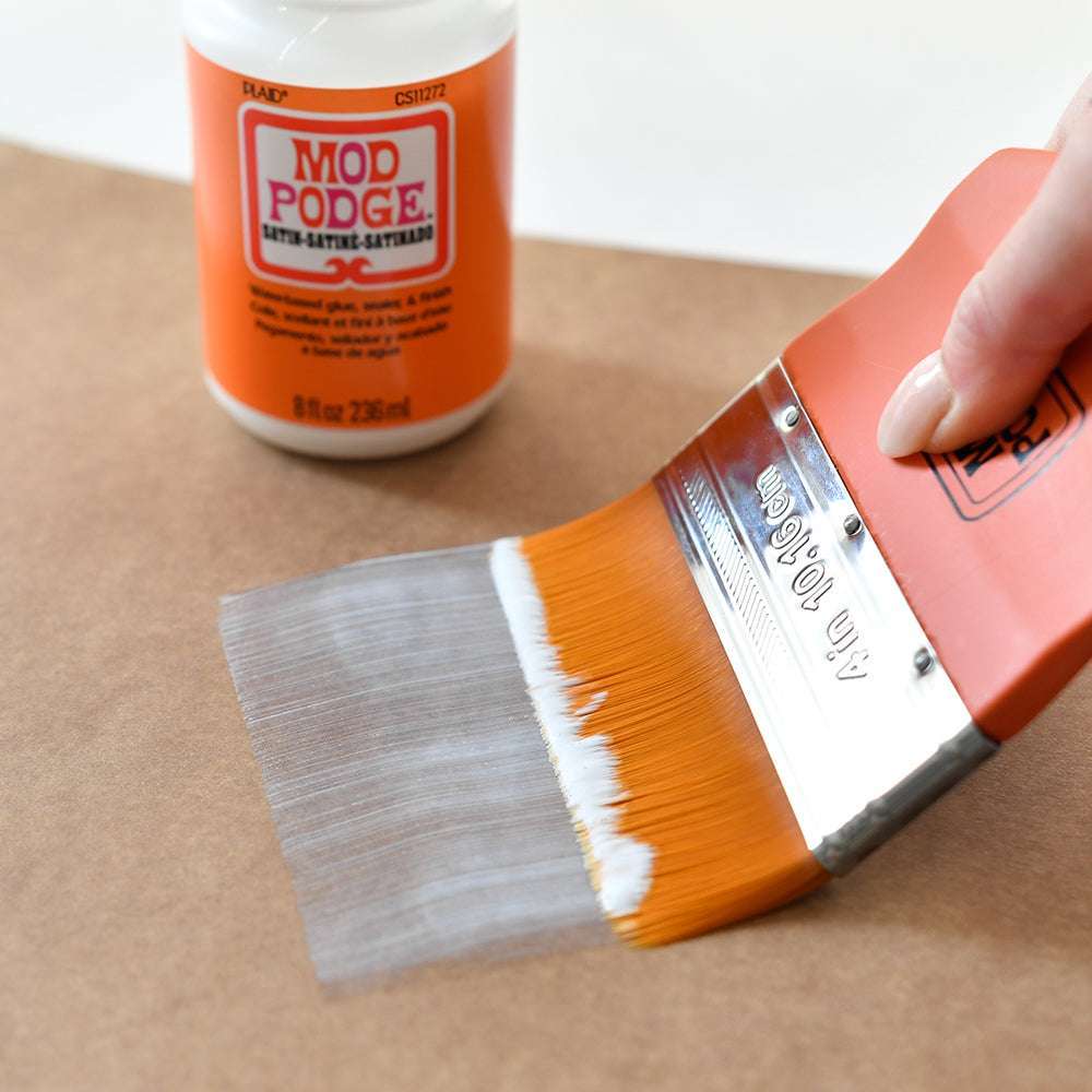 Eine Hand verwendet einen Plaid Enterprises Mod Podge-Pinsel mit Taklon-Borsten, um Mod Podge-Kleber auf braunes Papier für Decoupage-Projekte aufzutragen; im Hintergrund ist eine Flasche zu sehen.