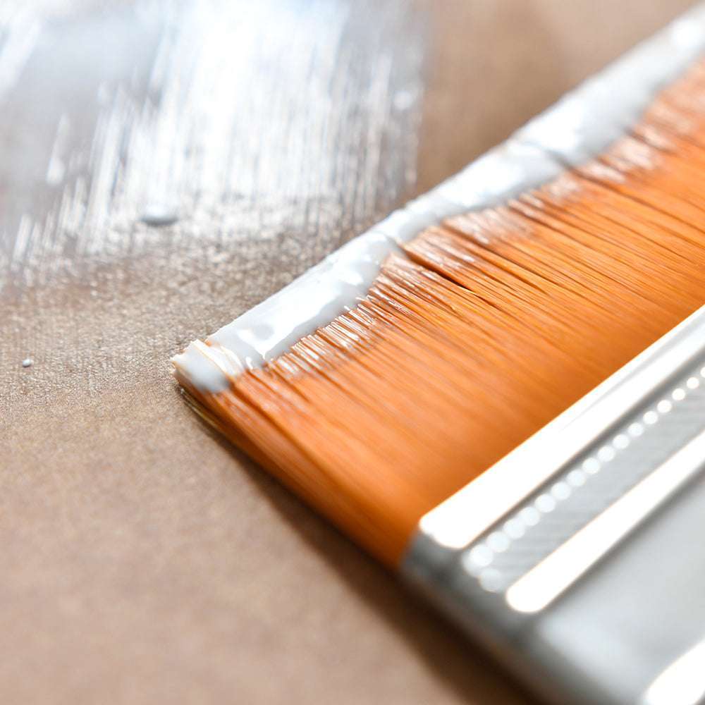 Nahaufnahme eines Plaid Enterprises Premium Borstenpinsels mit orangefarbenen Borsten, der weiße Farbe auf eine braune Oberfläche aufträgt.