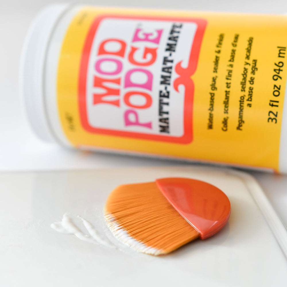 Eine Dose Plaid Enterprises Mod Podge und ein orangefarbener Decoupage-Pinsel mit Synthetikborsten ruhen auf einer weißen Fläche, ideal für Ihr nächstes Bastelwerkzeug-Projekt.