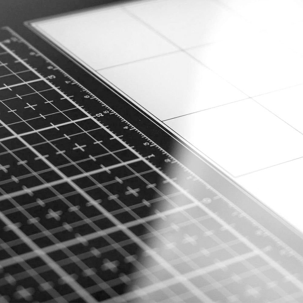 Eine Tonic Studios Glasmedienmatte mit Messrastern liegt unter hellem Licht neben einem leeren, geteilten Blatt Papier.
